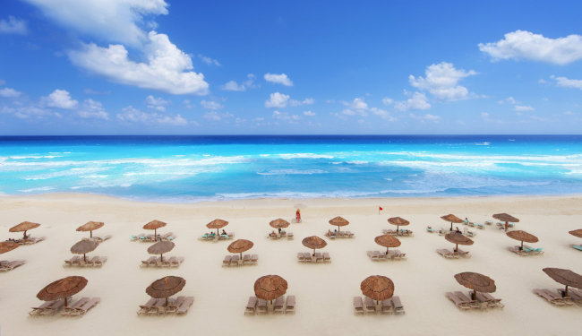 Aerial view of clear blue ocean with white sand, chairs with umbrellas in the foreground.